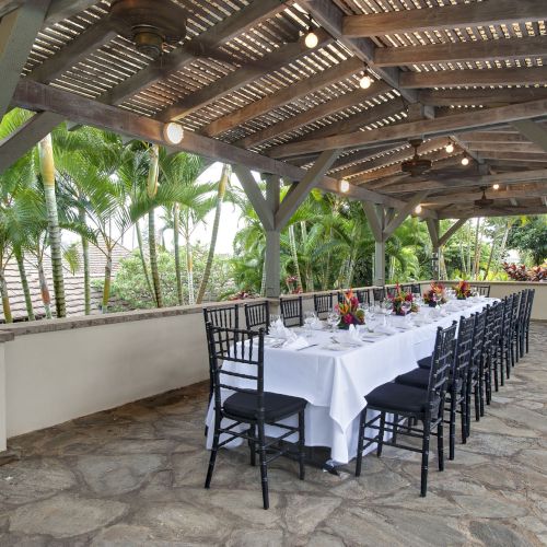 A long dining table set up outdoors under a wooden pergola with string lights, surrounded by lush tropical greenery and black chairs.