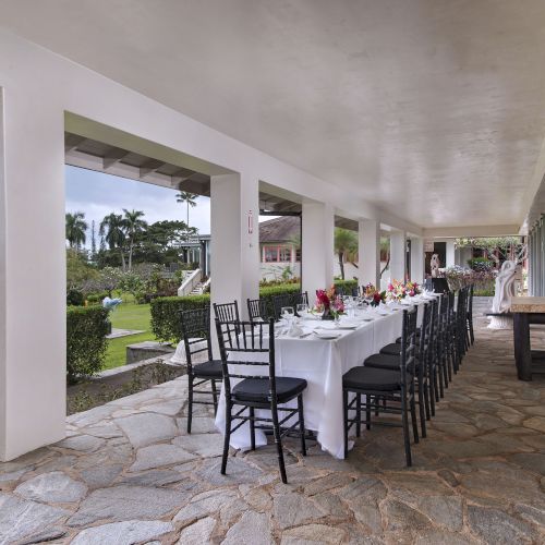 An outdoor dining setup with a long table, black chairs, and floral arrangements under a covered walkway, overlooking a garden.