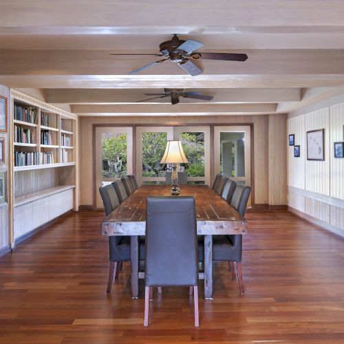 A spacious room with wooden floors features a long table with chairs, bookshelves, framed pictures, ceiling fans, and a lamp in the center ending the sentence.