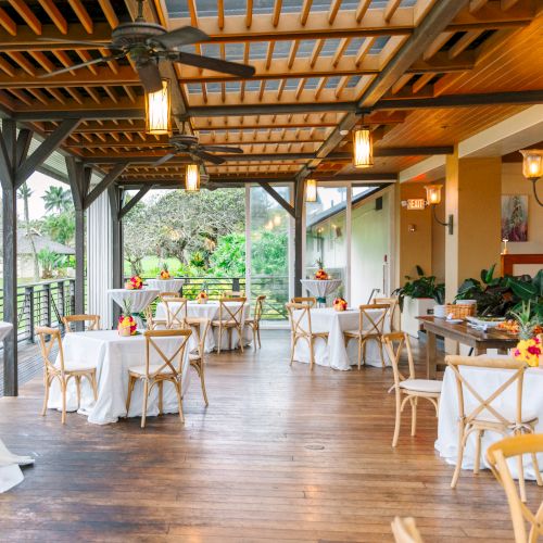 An elegant outdoor patio setup with decorated tables and chairs, ready for an event, overlooking a lush, green garden area.