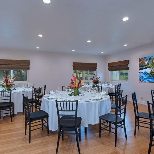 The image shows a banquet room with round tables set for dining, decorated with floral centerpieces, and surrounded by black chairs.