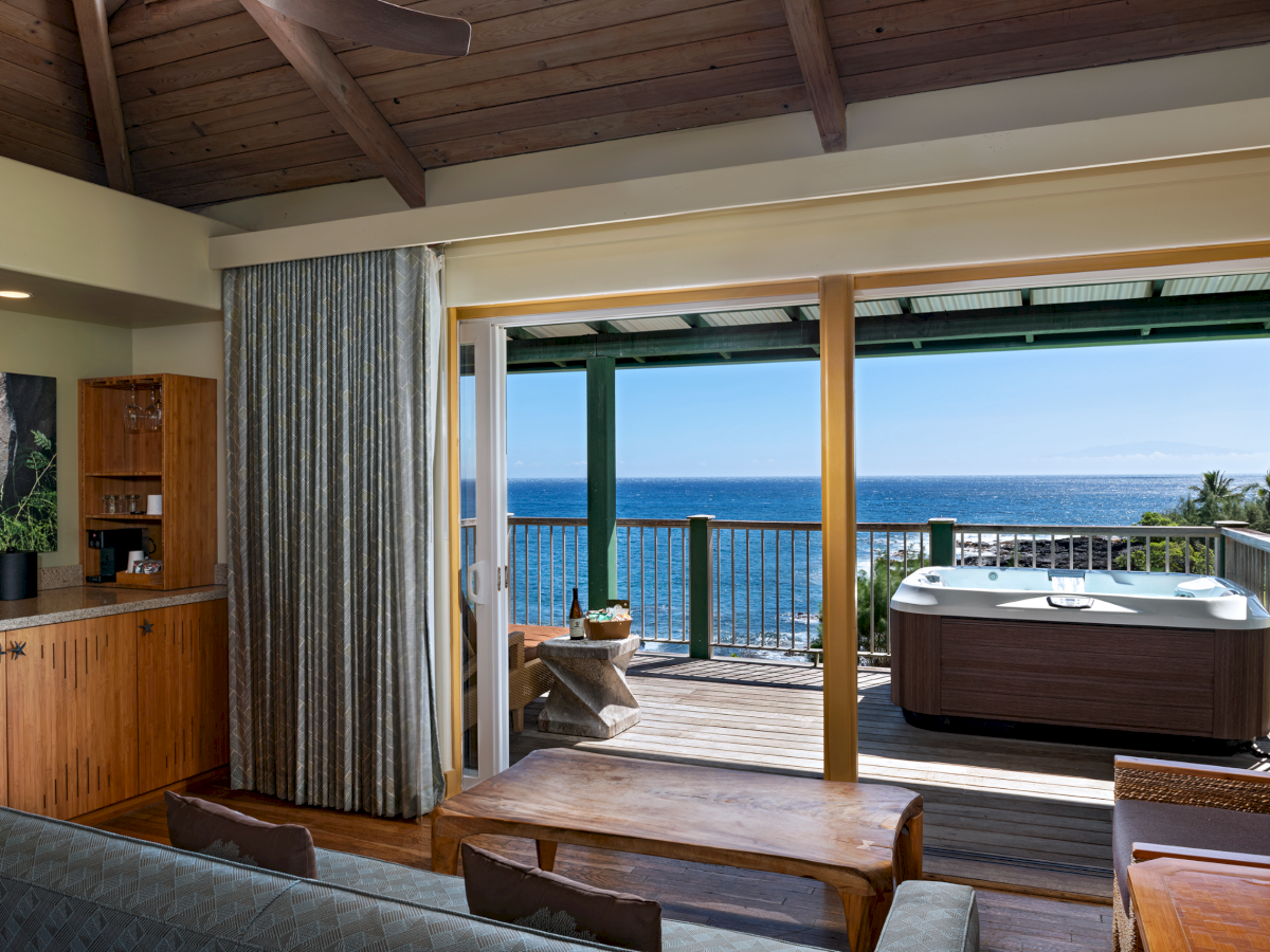 A cozy living room opens up to a deck with a hot tub overlooking the ocean under a clear blue sky.