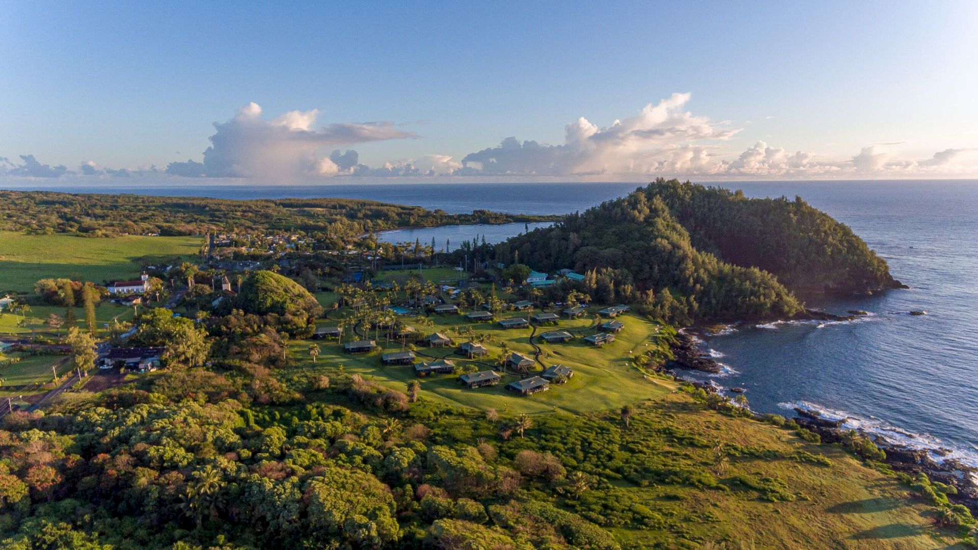 Aerial view of a coastal landscape with lush greenery, trees, scattered buildings, hills, and the ocean in the background under a partly cloudy sky.