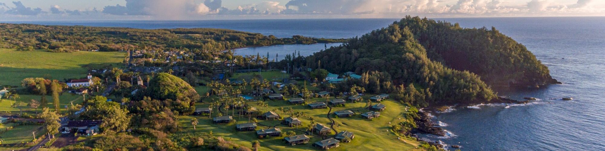 Aerial view of a coastal landscape with lush greenery, trees, scattered buildings, hills, and the ocean in the background under a partly cloudy sky.