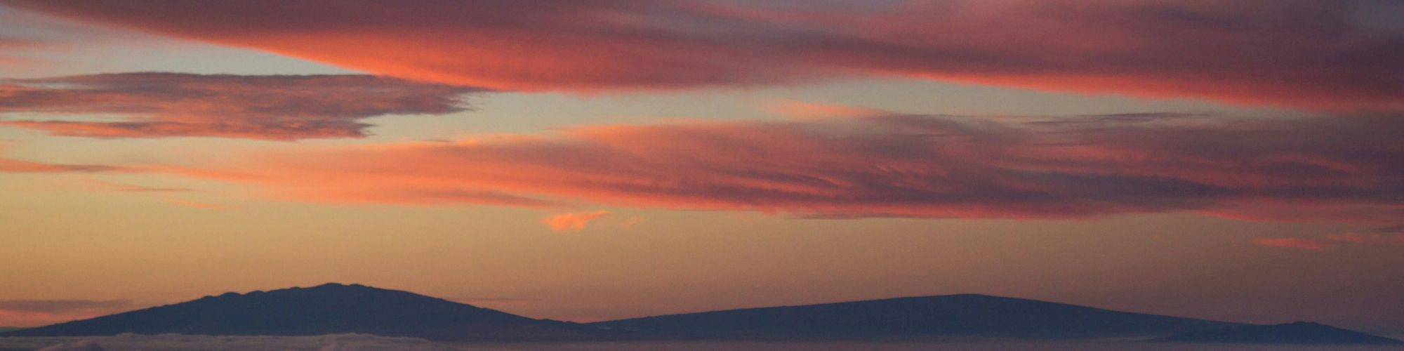 A serene landscape showing mountains silhouetted against a sky painted with soft pink and orange hues, with clouds hovering low in the horizon.