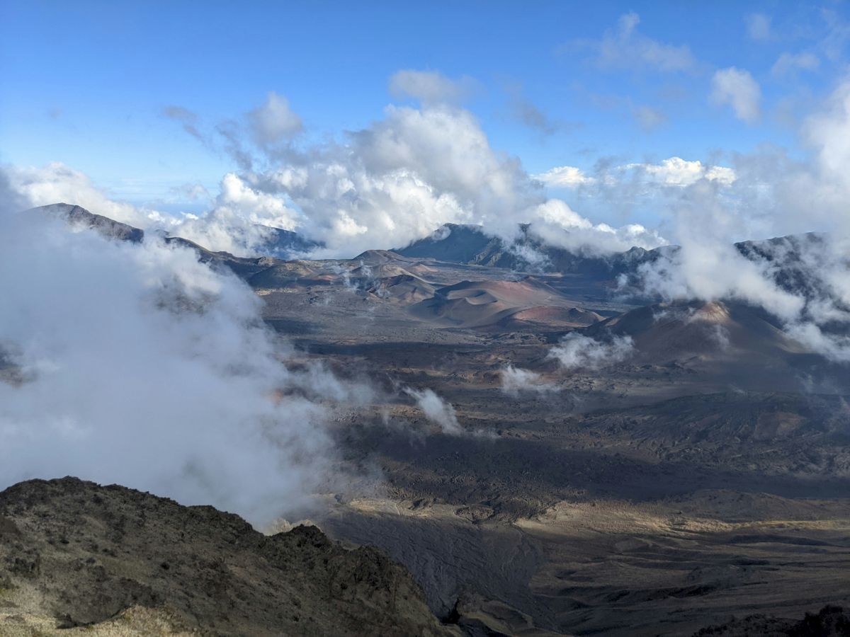 A scenic view of a mountainous landscape with clouds floating above, showing rugged terrain and distant peaks under a bright blue sky.