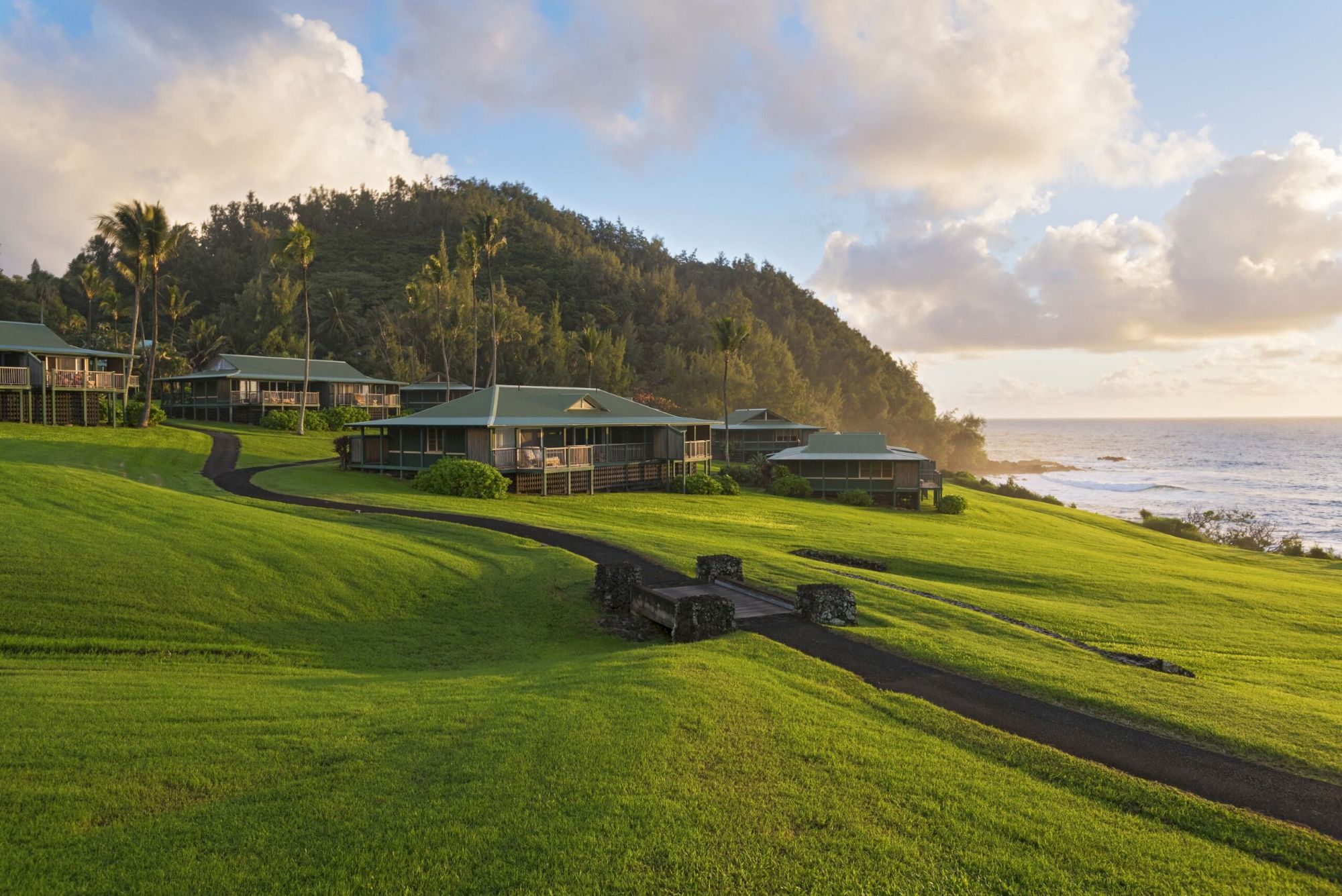 A scenic coastal landscape with lush green lawns, several buildings, a walking path, and a hill in the background under a partly cloudy sky, ending the sentence.