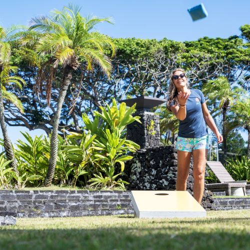 A person is playing cornhole outside, tossing a bean bag towards the board, surrounded by lush greenery and palm trees.