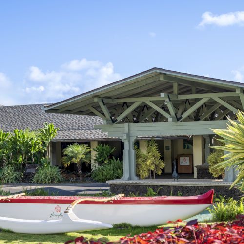 The image shows a building with a pitched roof, lush greenery, and a red-and-white canoe on the grass in front.