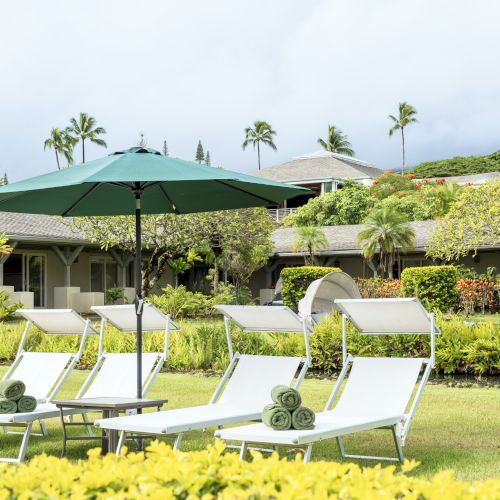 The image shows lounge chairs with rolled towels under an umbrella in a lush garden setting, with buildings and palm trees in the background.