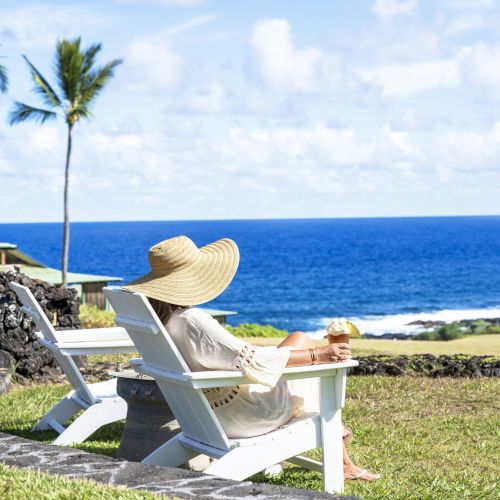 A person in a sun hat relaxes on a lounge chair, facing the ocean with palm trees and a house visible in the background.