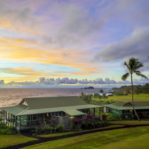 A coastal scene with a house, palm trees, and a vibrant sunset sky by the ocean, surrounded by lush green grass and colorful clouds.