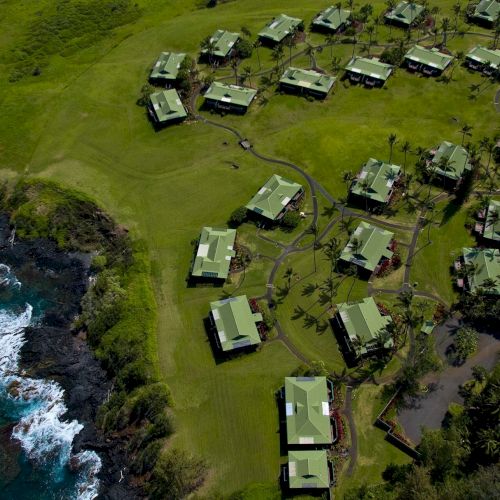 This image shows an aerial view of a coastal area with multiple buildings surrounded by green lawns and trees, near a rocky shoreline with waves.