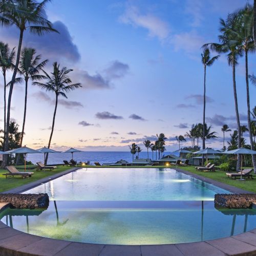 A luxurious outdoor pool surrounded by palm trees, deck chairs, and umbrellas, overlooking a scenic ocean view at sunset. The sky is partly cloudy.