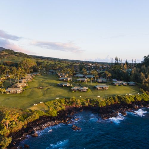 An aerial view of a coastal resort with multiple buildings surrounded by lush greenery, situated next to the ocean with a rocky shoreline.