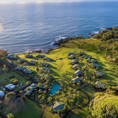 An aerial view of a coastal resort area with numerous buildings, lush greenery, and a swimming pool, all adjacent to the ocean and rugged shoreline.