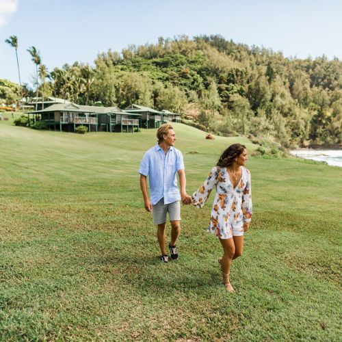 A couple is holding hands, walking on a grassy area near huts and a hilly landscape by the sea on a sunny day.