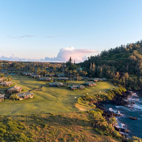 The image depicts a scenic coastal resort with cottages, lush greenery, hills, and ocean waves hitting a rocky shoreline under a clear sky.