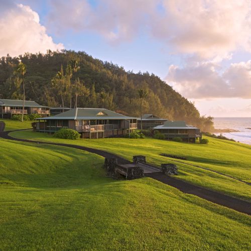 The image shows a scenic coastal landscape with green hills, several small buildings, a paved pathway, and the ocean in the background under a partly cloudy sky.