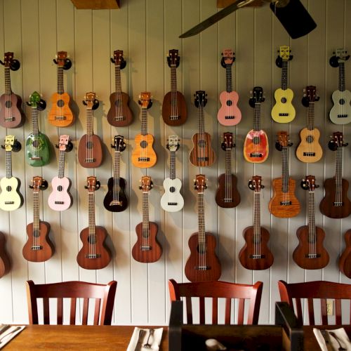 The image shows a wall with a colorful collection of ukuleles hanging neatly in rows above a wooden dining table and chairs.