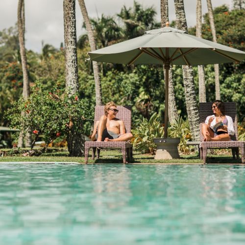 Two people are lounging on chairs beside a pool under an umbrella with lush greenery and trees in the background.