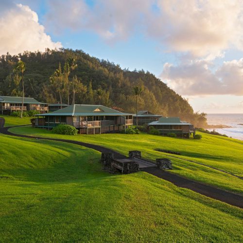 The image showcases a serene seaside landscape with green hills, palm trees, and cottages near the ocean, under a cloudy sky during sunset.