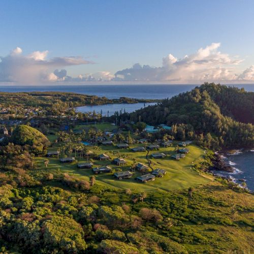 An aerial view of a coastal landscape featuring lush greenery, scattered houses, a forested hill, and the ocean extending to the horizon.