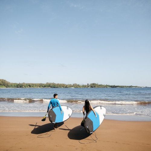 Two people walk towards the ocean on a sandy beach, carrying surfboards under a clear blue sky.