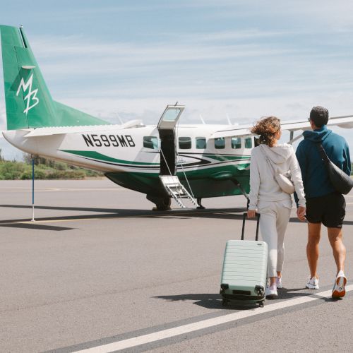 A couple walks toward a small green and white propeller airplane on a clear day, with one of them pulling a suitcase.