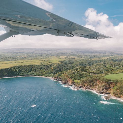 Aerial view of lush green coastline with cliffs and ocean waves, under an airplane wing, on a clear day.