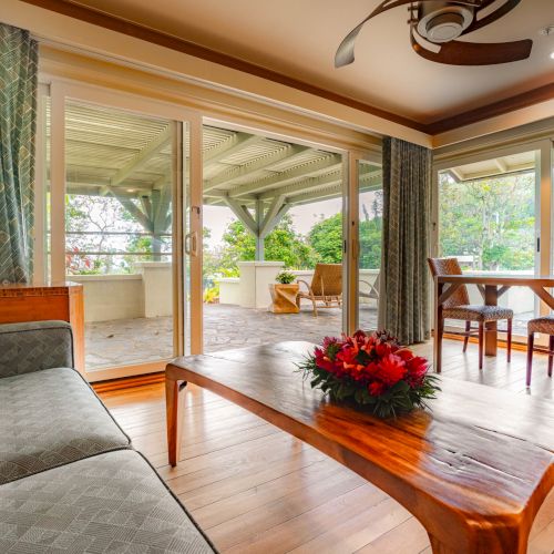 A cozy living space with a wooden coffee table, gray sofa, dining table, and chairs. Large windows open to a patio with outdoor seating and greenery.