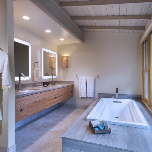 A modern bathroom with a bathtub, double sinks, large mirrors, wooden cabinets, and robes hanging on the wall. Natural light streams in through large windows.