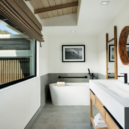 Modern bathroom with a freestanding bathtub, large window, twin sinks under round mirrors, and wooden ceiling accents.