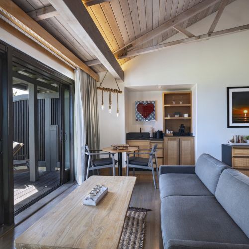 The image shows a modern living room with a grey couch, wooden coffee table, sliding glass doors, and a small kitchenette area with a framed picture.