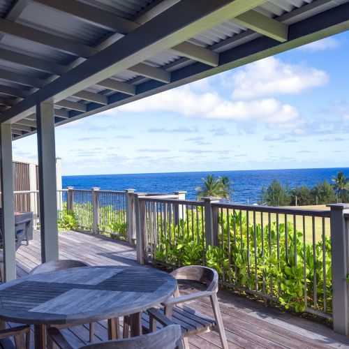 A wooden deck with seating overlooks the ocean, surrounded by greenery under a partially sunny sky, offering a serene coastal view.