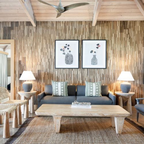 A modern living room featuring a wooden theme, with a sofa, chairs, coffee table, and two framed artworks above, illuminated by table lamps.