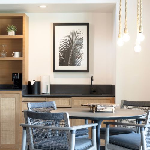 A modern kitchenette with a dining area features a round table, wicker chairs, a sink, and shelves with decor, plants, and kitchenware.