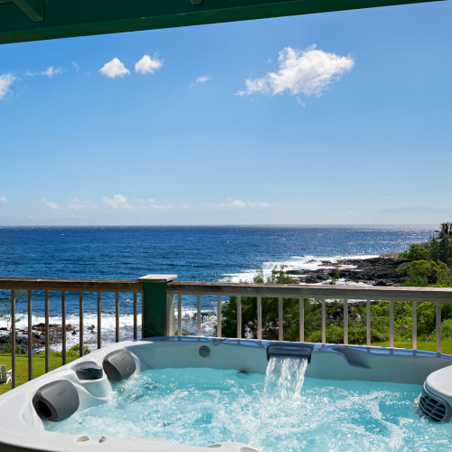 A hot tub on a deck overlooks the ocean with a stunning coastal and grassy landscape in the background, under a partly cloudy blue sky.