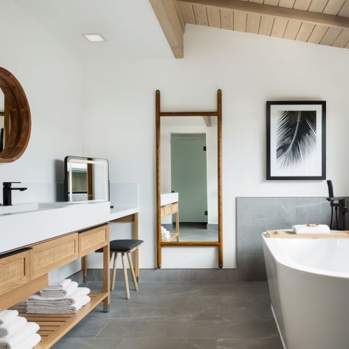 A modern bathroom with a double sink, round mirrors, wooden accents, a free-standing bathtub, a tall mirror, and a framed leaf artwork on the wall.