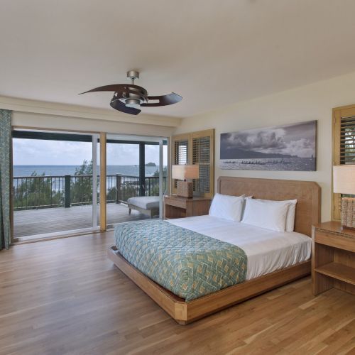 A spacious bedroom with a large bed, wood flooring, nightstands, lamps, and a ceiling fan, overlooking a balcony with a sea view.