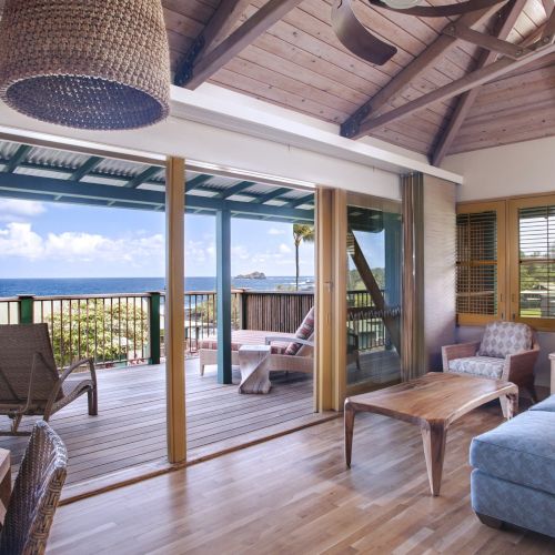 A cozy living room with wooden furniture, large windows, and a view of the ocean. Sliding doors open to a balcony with lounge chairs.