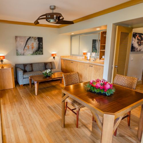 A cozy, well-lit living and dining area with wooden flooring, a dining table adorned with flowers, and a sofa against the back wall.