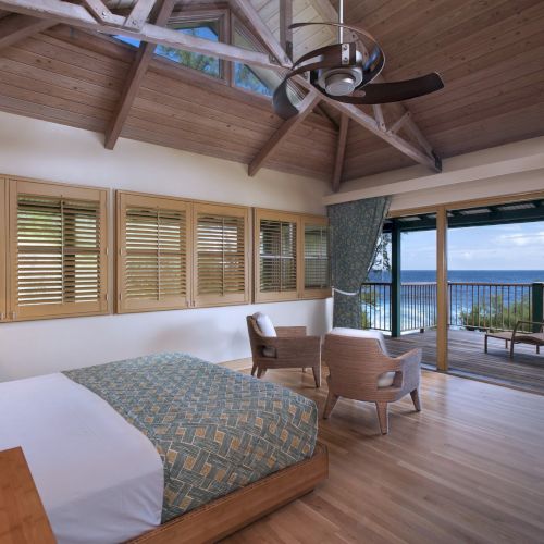 A bedroom with wooden floors, vaulted ceiling, and large windows opens to a balcony with ocean views. Two chairs and a bed are visible.