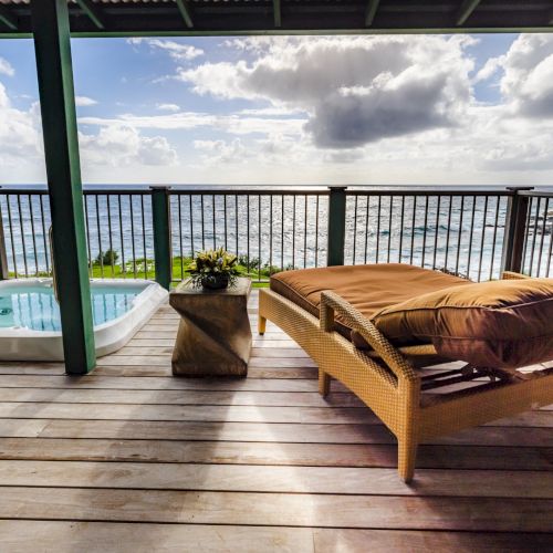 A serene deck overlooks the ocean, featuring a hot tub, cushioned loungers, and a small table, all under a covered terrace with a stunning view.