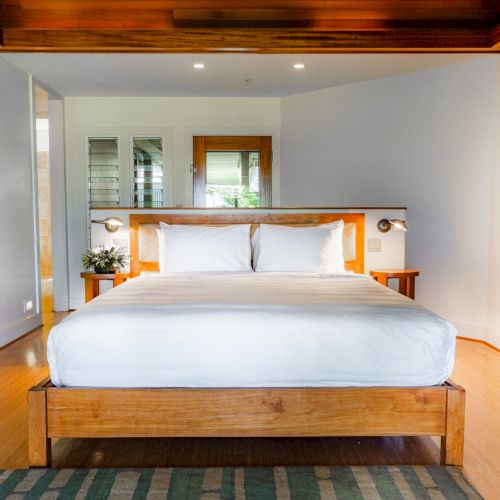 The image shows a modern bedroom with a wooden bed, white linens, a wooden ceiling, and a painting on the wall, all in a bright setting.