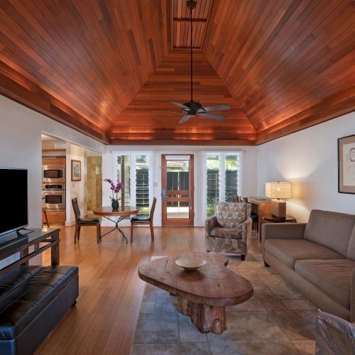 A cozy living room with wooden ceiling, TV, sofa, armchairs, coffee table, wall art, and windows leading to another space, featuring warm lighting.