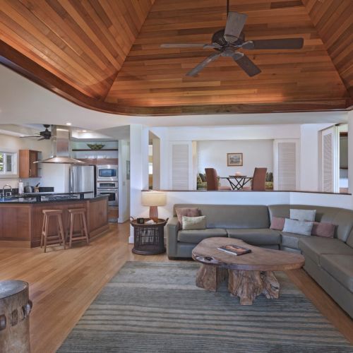 The image features a spacious living room with a large L-shaped sofa, a wooden coffee table, an open kitchen, and a ceiling fan, ending the sentence.