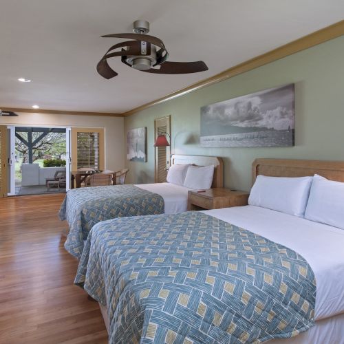 A spacious hotel room features two double beds, a ceiling fan, wooden accents, and a seating area with a view of the outdoors through a large window.