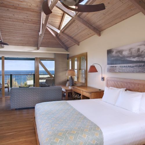 A cozy bedroom with wooden decor, a ceiling fan, and a comfortable bed. There is a seating area and a balcony with a view of the ocean.