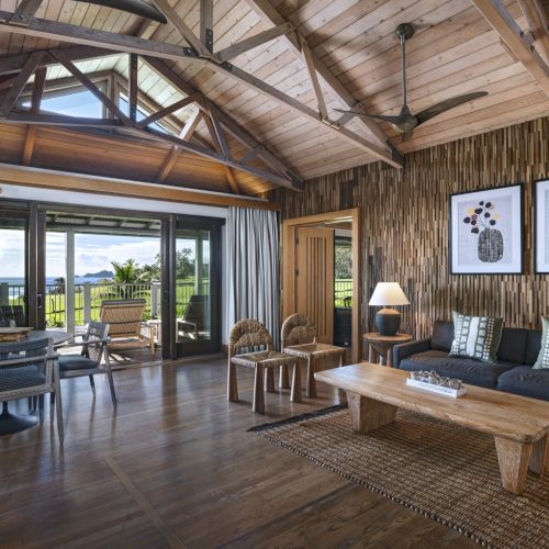 A cozy living room with wooden accents, a sofa, coffee table, wall art, and large windows opening to a scenic view, featuring natural light and decor.